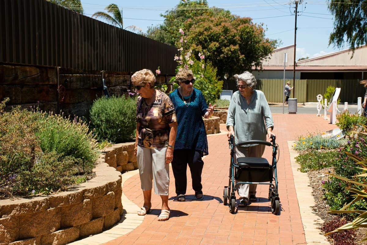 Three older women walking together along a sunny garden path, one using a rollator