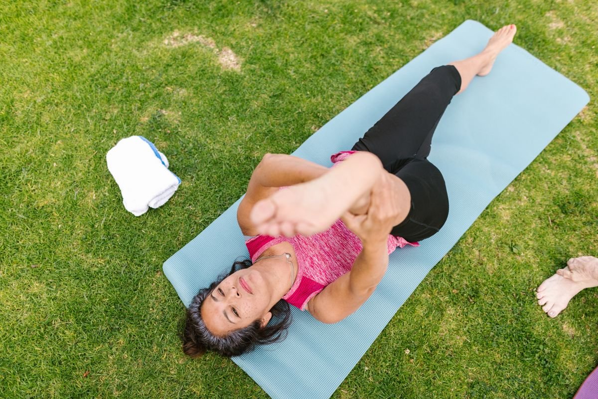 Mature woman lying on a yoga mat outdoors gently pulling one knee toward her chest for a leg stretch